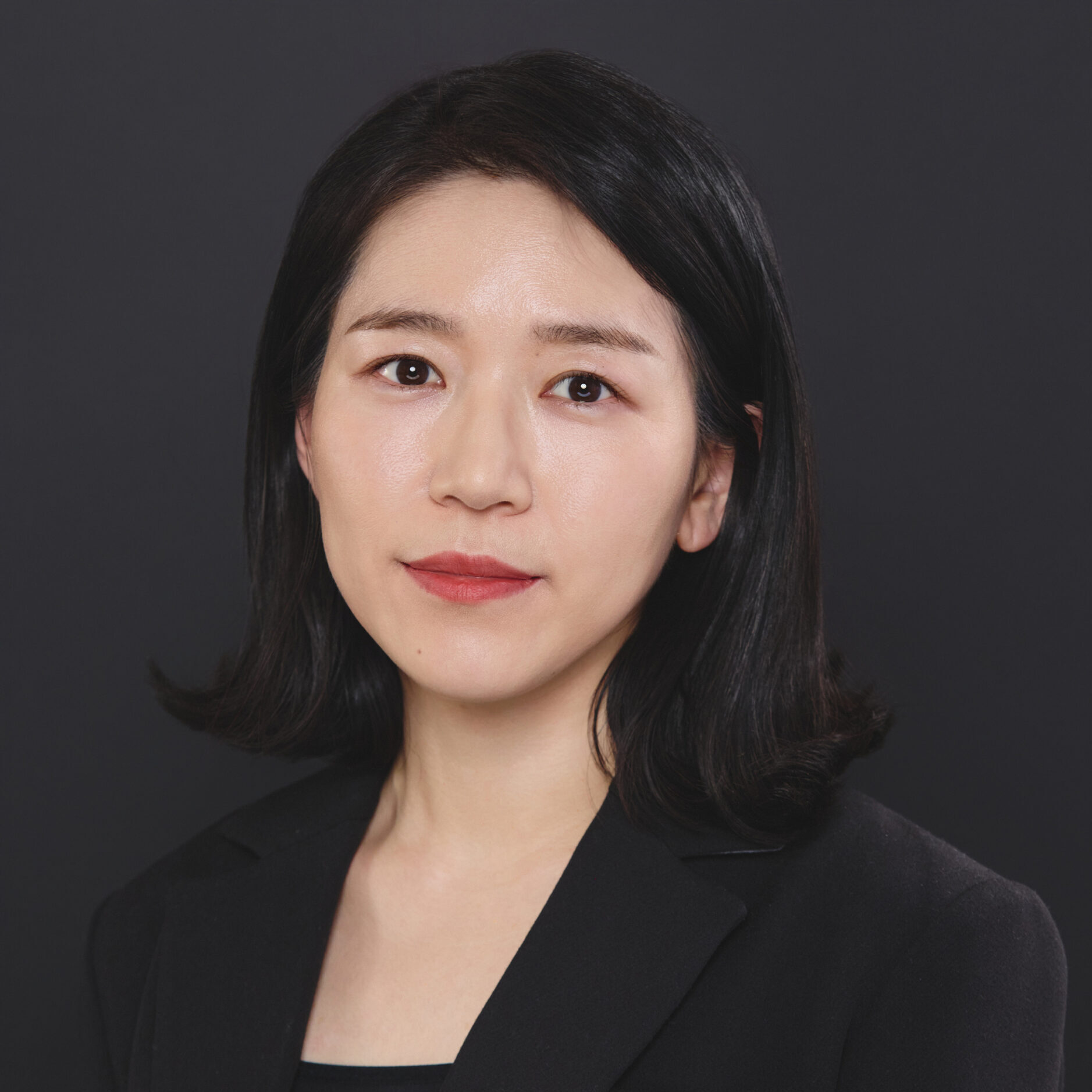 A woman with straight black hair, wearing a black blazer, poses against a plain dark background.
