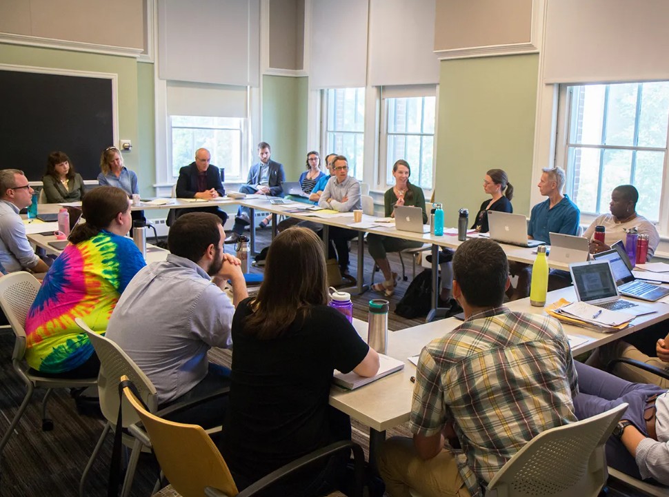 People sit around a large table in a well-lit meeting room, engaged in discussion, with laptops, notebooks, and water bottles in front of them.