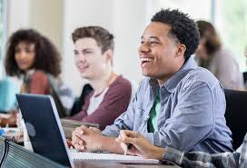 A group of students sit at desks in a classroom, with notebooks and a laptop, listening and smiling during a lesson.