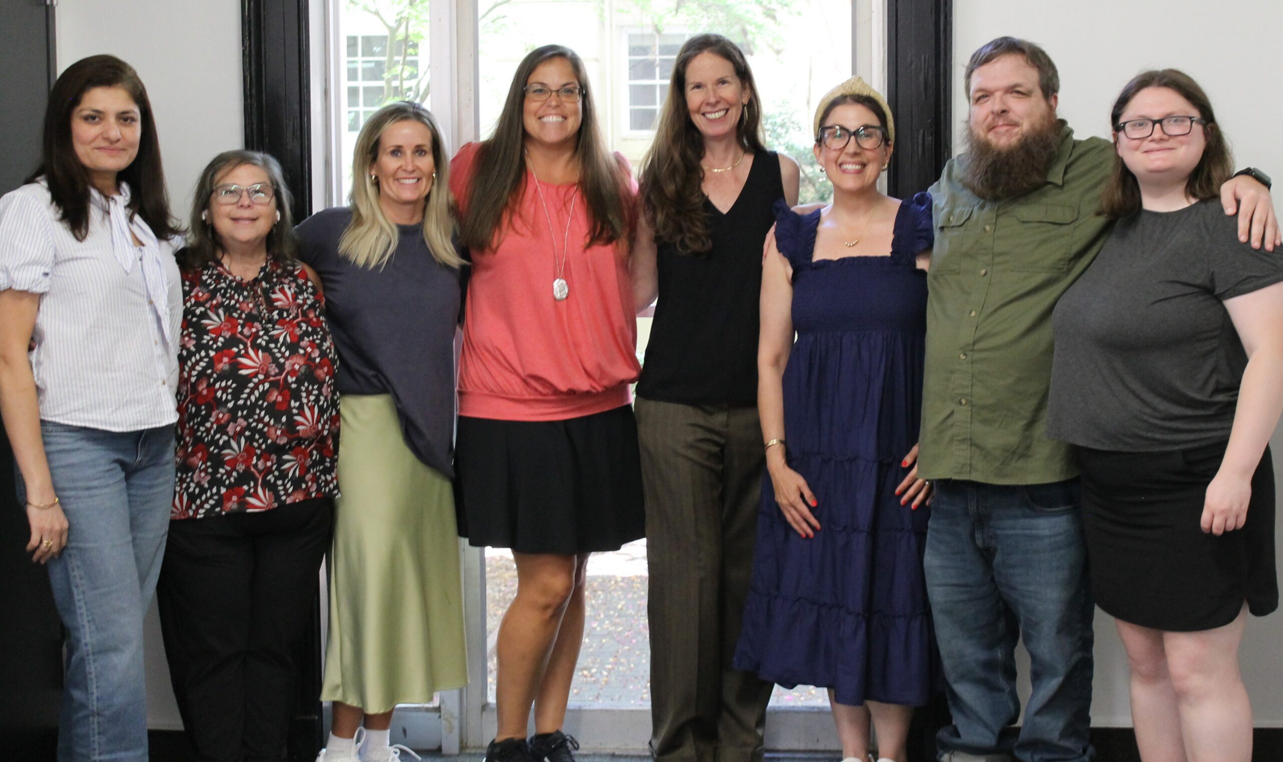 Eight staff members from the Owens Institute stand smiling side by side indoors, with a window and outdoor greenery visible in the background.